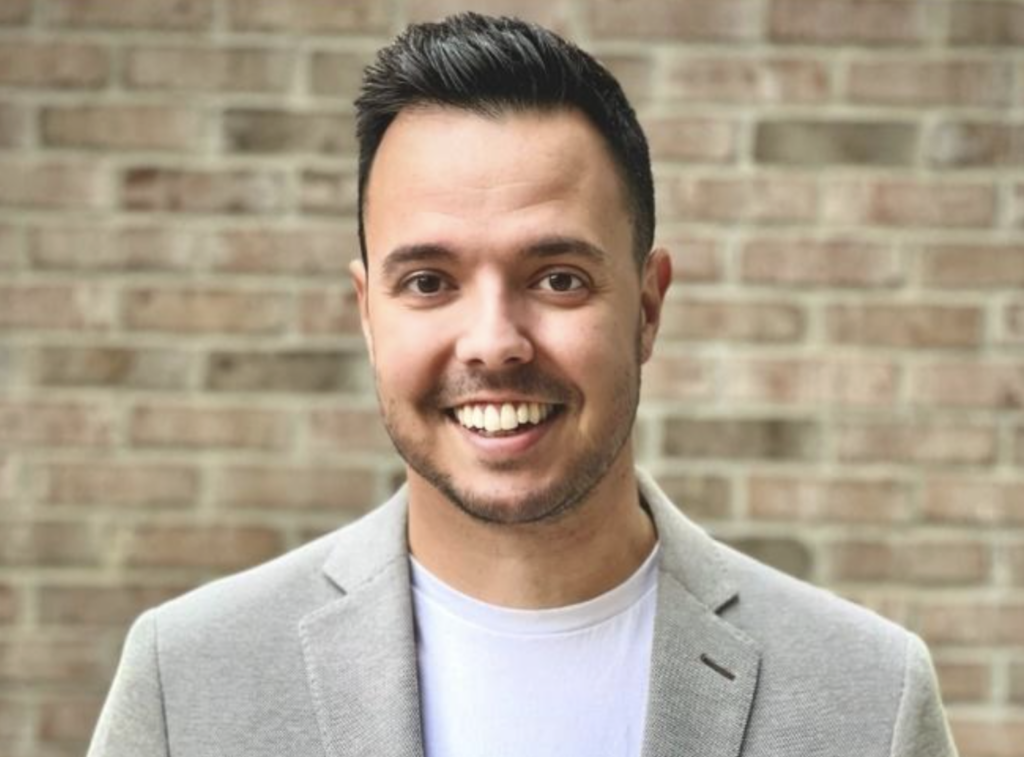 A smiling person in a light gray blazer and white shirt stands in front of a brick wall.